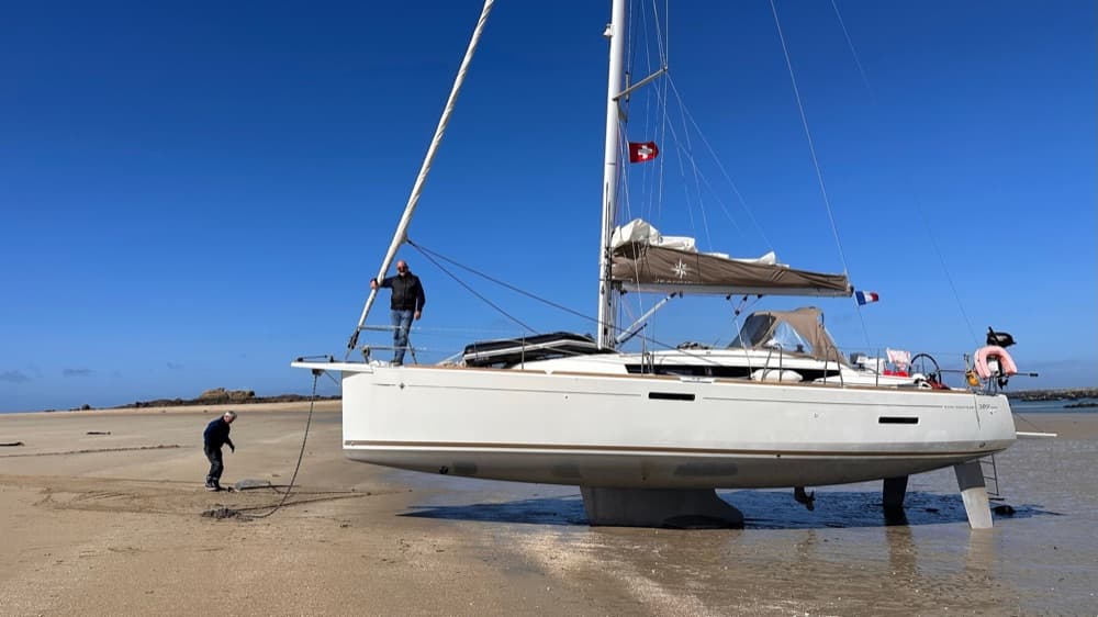 Segelyacht mit Schweizer Flagge bei Ebbe trockengefallen auf einem Sandstrand, Crew an Deck und am Strand