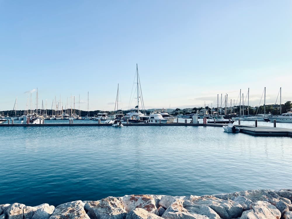 Hafen mit Segelyachten an den Stegen, ruhiges Wasser und Steinmole im Vordergrund