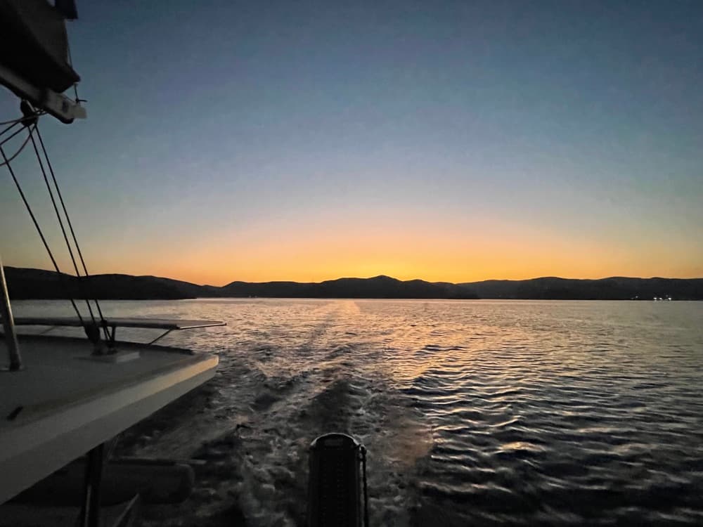Blick vom Heck einer Yacht aufs Kielwasser in der Abenddämmerung, am Horizont eine Hügelkette