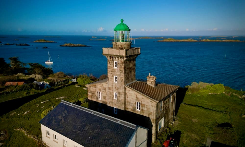 Leuchtturm aus Granit mit grüner Laterne auf einer Insel, dahinter ankernde Segelboote zwischen Felsinseln
