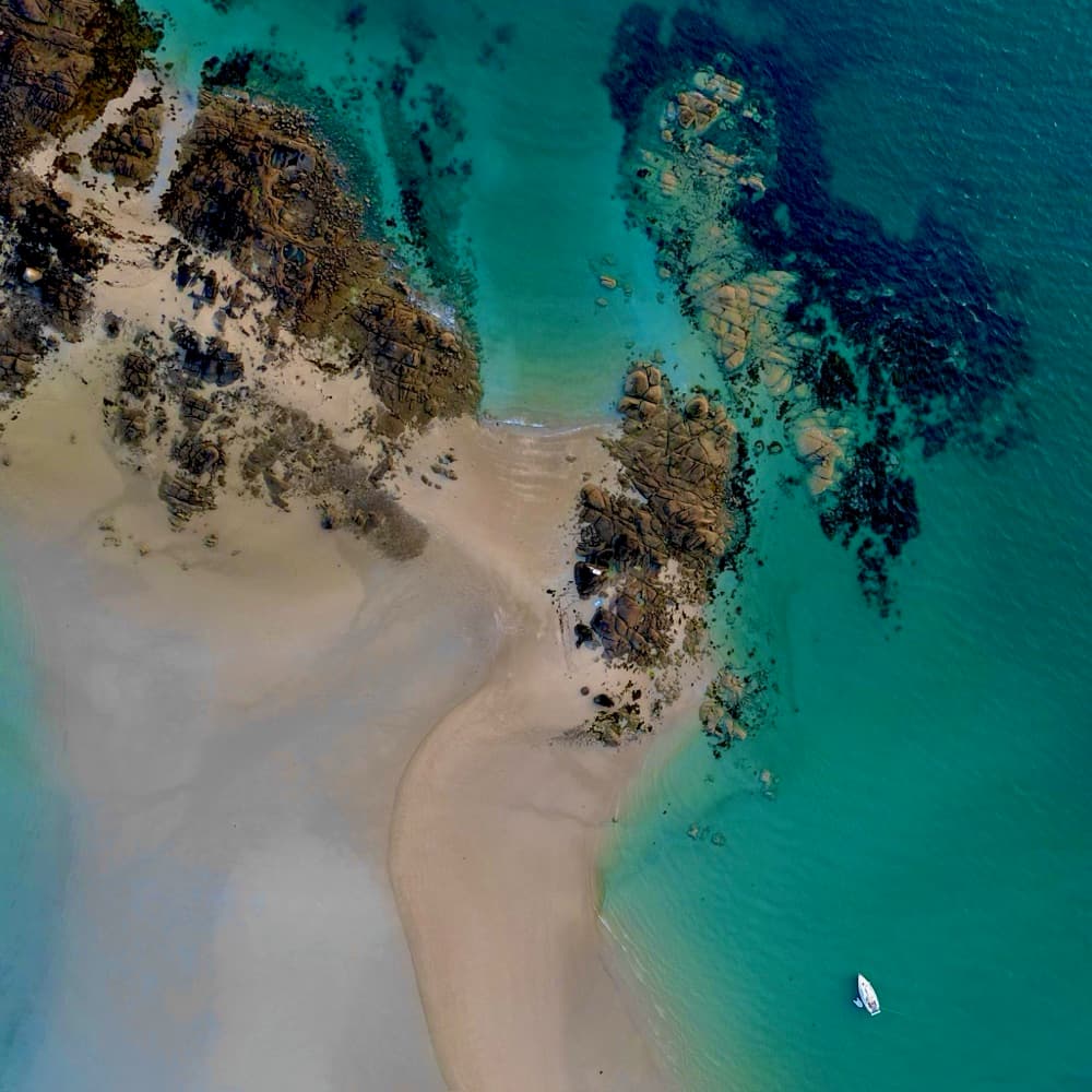 Luftaufnahme von oben: Sandbank, Felsen und türkisfarbenes Wasser, daneben ein ankerndes Boot
