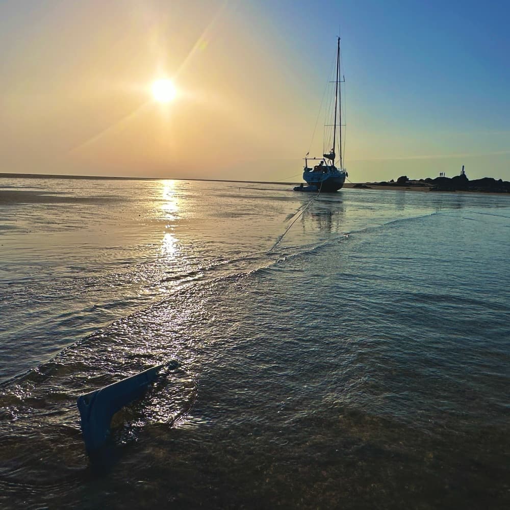 Anker im flachen Wasser, dahinter eine Segelyacht im Gegenlicht der Abendsonne
